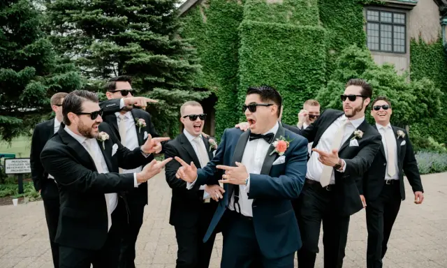 Groomsmen in black suits and cream ties, with the groom in a navy suit, all sporting sunglasses and a playful mood. Do groomsmen match the groom? Not exactly, but they're all dressed to impress!