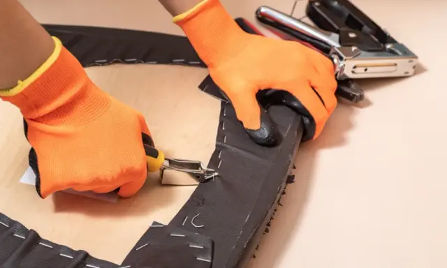 Manos con guantes naranjas usando una grapadora para tapizar una silla.