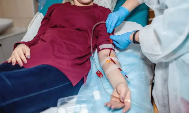 Pacjentka leży, a pielęgniarka w rękawiczkach podłącza przetokę do dializ. Krew płynie przez rurki.