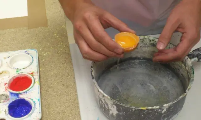 Manos vierten una yema de huevo en un recipiente para preparar una pintura al temple. Paleta con pigmentos de colores al lado.