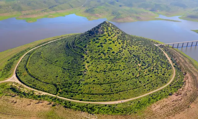 El **cerro Masatrigo**, cubierto de vegetación, se alza majestuoso junto a un embalse. Una carretera serpentea a su alrededor, conectando con un puente.