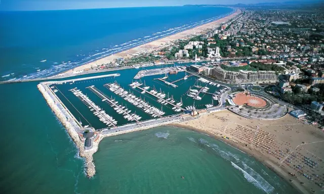 La darsena di Rimini, con le sue barche ormeggiate, si estende lungo la costa adriatica, tra la spiaggia affollata e il mare azzurro.