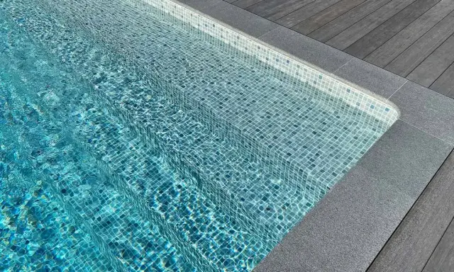 Trappsteg i en pool med blå mosaik, kantade av grå sten och trätrall. Sarg runt poolen ger en lyxig känsla.