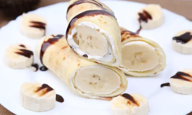 Rolowane naleśniki z bananem i kremem, polane czekoladą. Na talerzu ułożone są też kawałki banana z polewą.
