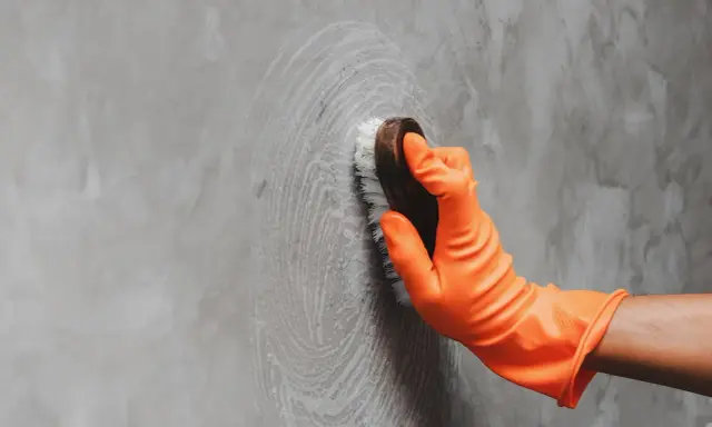 Mano con guante naranja limpiando pared gris con cepillo, aprendiendo como limpiar las paredes de color sin dañar la pintura.