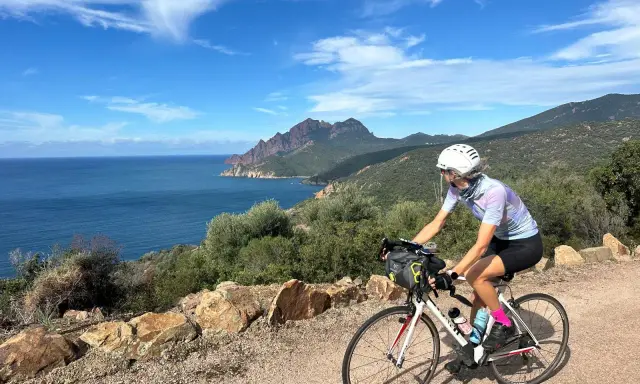 Ciclista pedala lungo la costa rocciosa della Corsica, con il mare blu e montagne sullo sfondo. Un'avventura in bici indimenticabile.