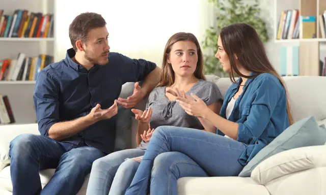 Tres personas sentadas en un sofá blanco discuten animadamente, gesticulando con las manos, en un intento por resolver conflictos.