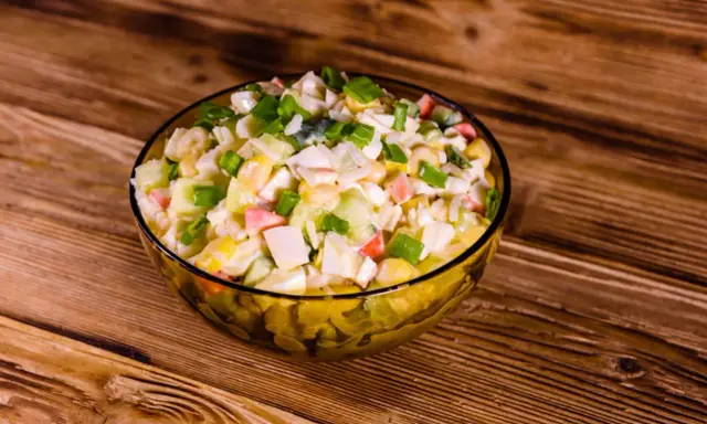 Pyszna sałatka z surimi, z kukurydzą, jajkiem i szczypiorkiem, podana w szklanej misce na drewnianym blacie.