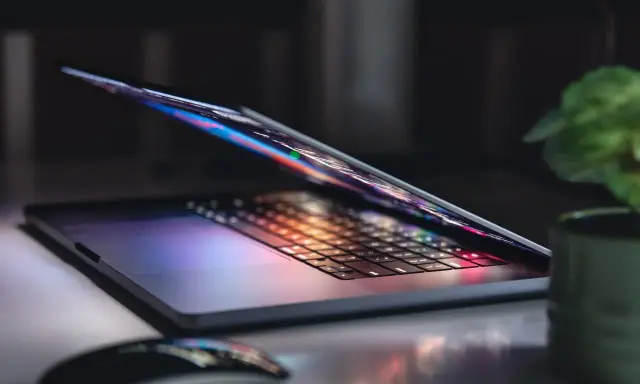 Otwarty notebook z podświetlaną klawiaturą i ekranem, obok myszka i roślina. To jest właśnie notebook, narzędzie do pracy i rozrywki.