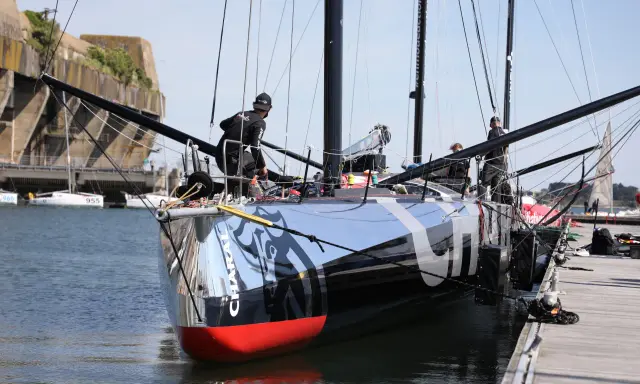 L'IMOCA Charal, un voilier de course futuriste, est amarré à quai. Des marins s'affairent sur le pont, préparant le navire pour une nouvelle aventure en mer.