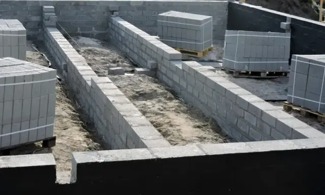 Budowa domu w toku: wykopy pod fundamenty i pierwsze ściany z bloczków betonowych. Materiały budowlane czekają na użycie.