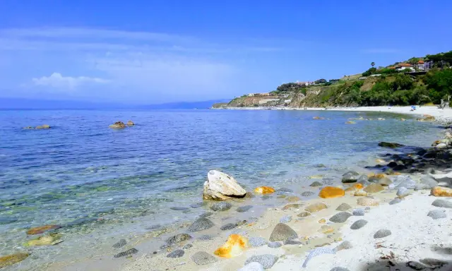 Spiaggia di Tropea con la torre Sant'Irene in lontananza, mare cristallino e rocce sulla riva.