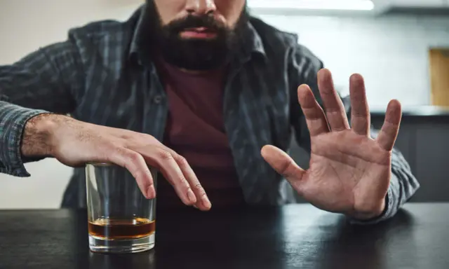Mężczyzna z zarostem odpycha szklankę z alkoholem, walcząc z chorobą alkoholową. Gest dłoni symbolizuje odmowę i chęć zmiany.