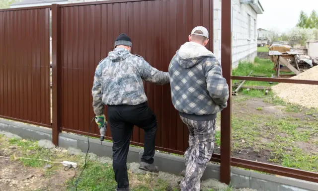Dwóch mężczyzn montuje brązowy płot. Jeden z nich trzyma wiertarkę. Czy można postawić płot przy płocie?