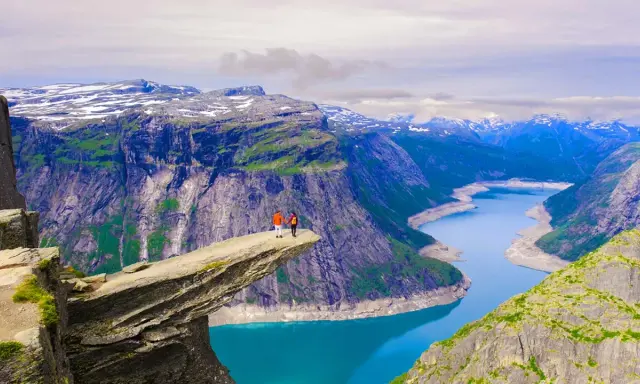 Två personer står på Trolltunga, en av Norges mest ikoniska och fina platser. En dramatisk klippavsats med en turkos sjö nedanför och snöklädda berg i bakgrunden.