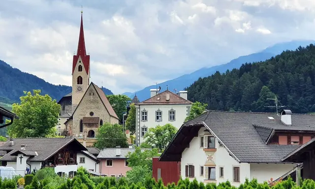 Un borgo pittoresco in Val d'Isarco, con una chiesa dal campanile rosso svettante tra case tradizionali e montagne boscose.
