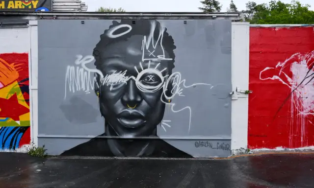 En imponerande **graffiti** av ett ansikte med en krona och solstrålar runt ögonen. Konstverket är målat i gråskala med vita detaljer.