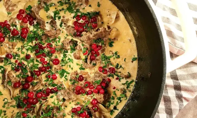 En närbild på en krämig renskavsgryta med färska lingon och persilja, serverad i en gjutjärnspanna.