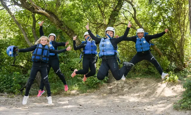 Cinco personas, incluyendo niños, saltan alegres con trajes de neopreno y chalecos salvavidas en un entorno natural, listos para una aventura en Huesca.