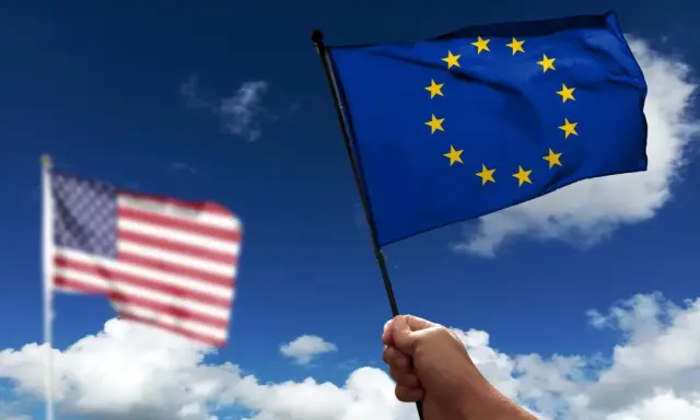 Hand holds EU flag, US flag blurred in background. Discussing what is soevereiniteit in a global context.