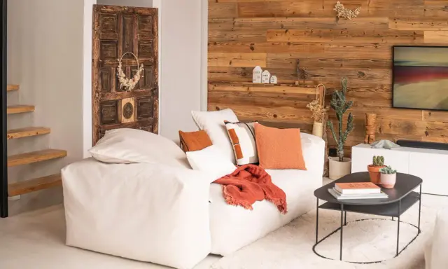 Un salon chaleureux et accueillant avec un grand canapé blanc, des coussins orange et un mur en bois rustique. Une porte ancienne décore le mur.