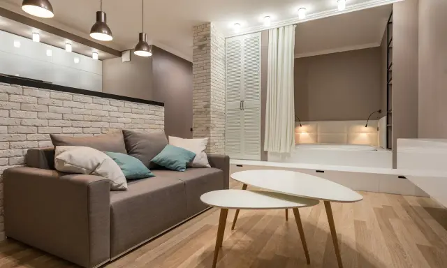 Nowoczesny salon z sofą i stolikami kawowymi. W tle widoczna ściana ze starej cegły, łóżko i szafa.