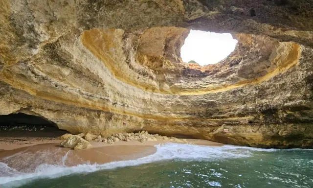 Die Benagil-Höhle mit ihrem kreisförmigen Loch im Felsdach, das Sonnenlicht hereinlässt. Wellen brechen sanft am Sandstrand im Inneren.