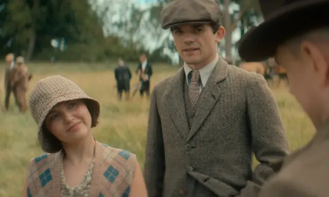 Giovane donna con cappello a scacchi e gilet a rombi, accanto a un uomo con berretto e giacca di tweed. Sullo sfondo, i sette quadranti di un campo.