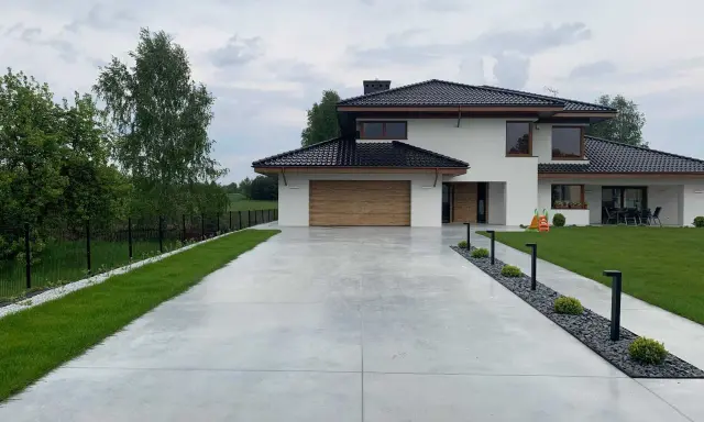 Nowoczesny dom z drewnianą bramą garażową, otoczony zielenią. Szeroki podjazd betonowy prowadzi do wejścia, a wzdłuż niego biegną lampy i rabaty z kamieniami.