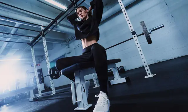 Donna in tenuta sportiva nera, con scarpe bianche, seduta su panca in palestra. Ispirazione su come vestirsi in palestra.