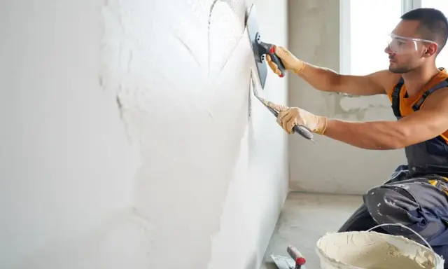 Un ouvrier, protégé par des lunettes et des gants, s'applique à crépire un mur avec une truelle.