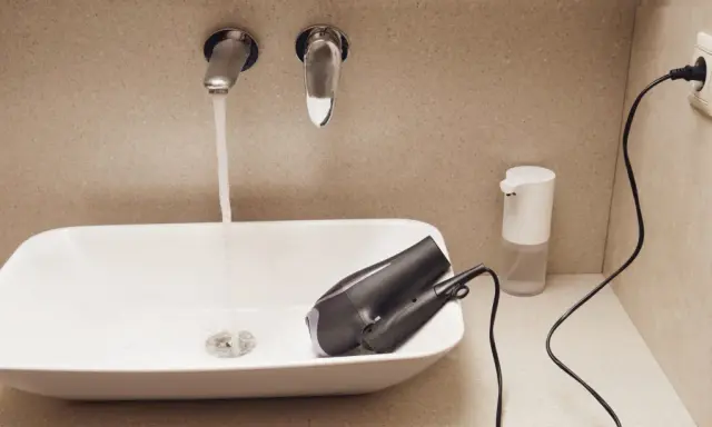 Prise salle de bain : un sèche-cheveux est posé dans un lavabo où coule l'eau. Un distributeur de savon et une prise électrique sont visibles.