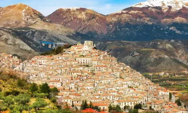 Un pittoresco borgo calabrese si aggrappa a una collina, con case in pietra e tetti rossi che si estendono fino alle montagne innevate.