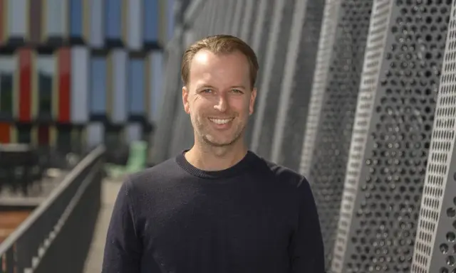 Daniel Larsson, skådespelare, ler mot kameran. Bakgrunden är en modern byggnad med geometriska mönster.