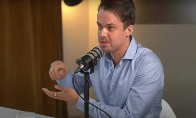 Michael van de Poppe w niebieskiej koszuli mówi do mikrofonu, gestykulując.