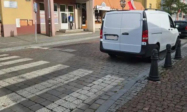 Biały van blokuje przejście dla pieszych na brukowanej ulicy.