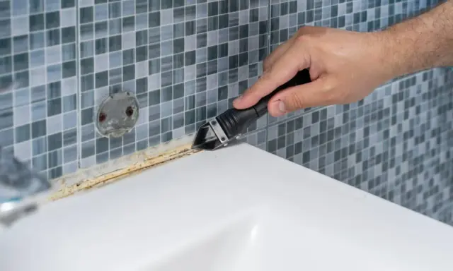 Mano usando una espátula para quitar silicona vieja y sucia de la bañera.