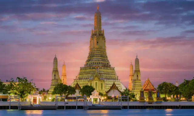 Il **tempio dell'alba Bangkok** (Wat Arun) si erge maestoso al tramonto, con le sue guglie dorate che brillano contro un cielo rosa e viola.