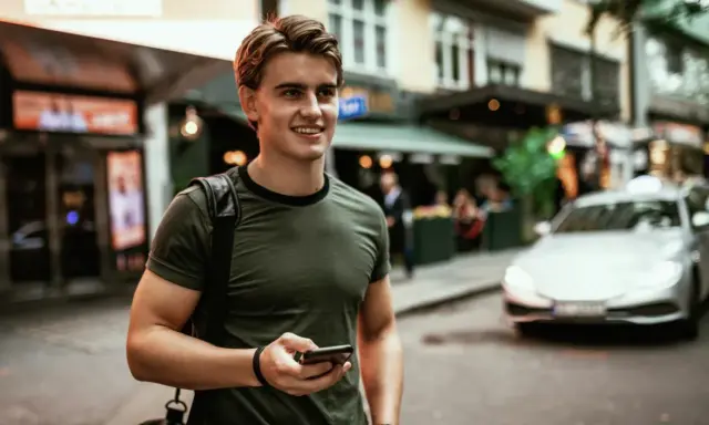 Ung man med mobiltelefon i handen, på en stadsgata med bilar och butiker. Känns som vårt hem utomlands.