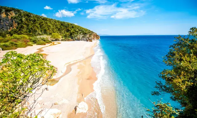 En orörd albanien strand med turkost vatten, vit sand och gröna kullar. En liten flod rinner ut i havet.