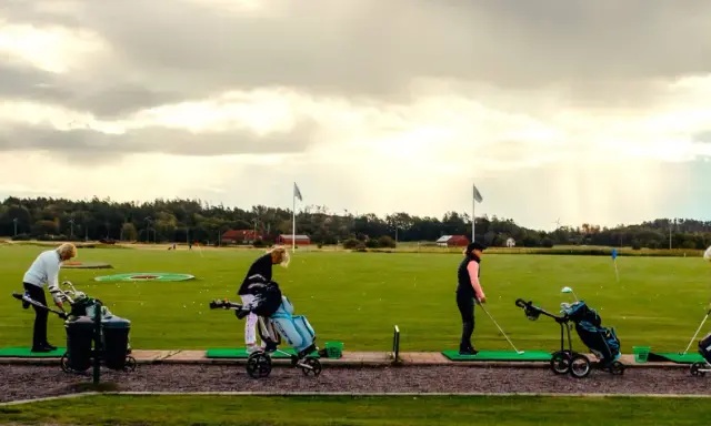 Kvinnor tränar golf på en driving range i Sverige. Solstrålar bryter igenom molnen.