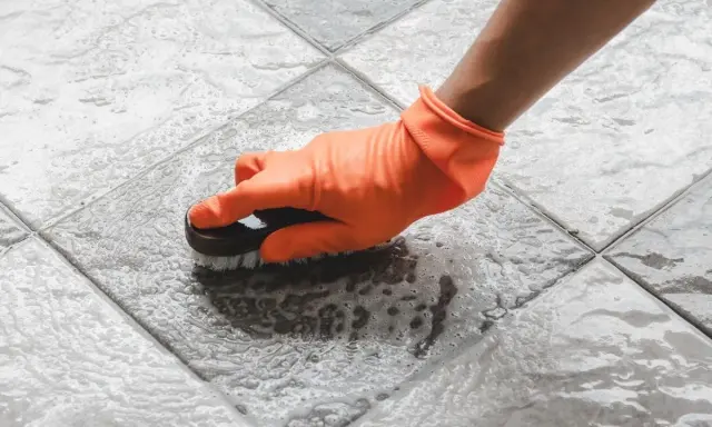 Mano con guante naranja cepillando un suelo antideslizante con espuma.