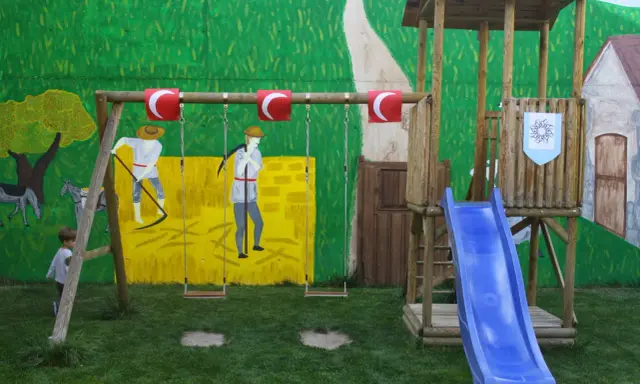 Un niño observa un columpio y un tobogán azul, parte de unos juegos medievales para niños, con un mural de fondo.