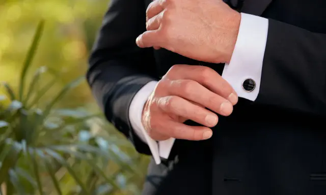 A man fastens his black cufflinks on his white shirt cuffs, completing his tux.