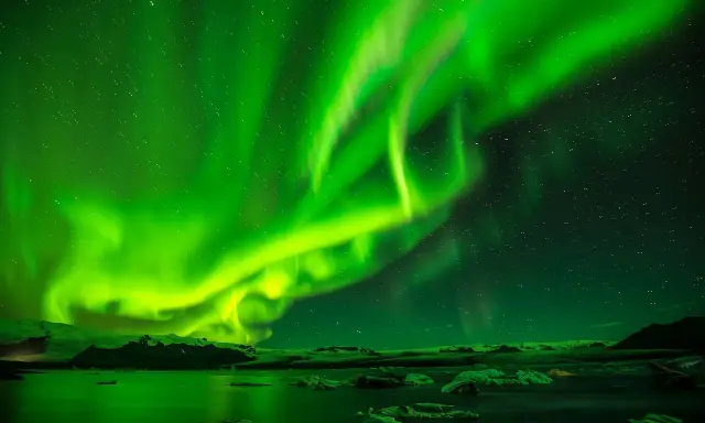 Grön norrsken dansar över en isig ö med stjärnor som glittrar. Vattnet speglar det magiska skådespelet.
