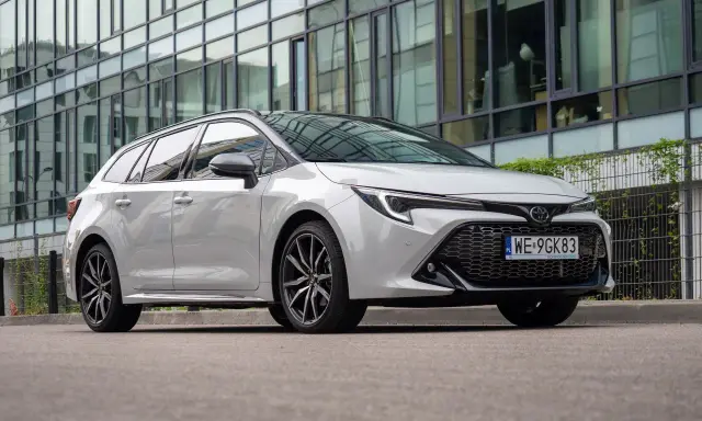 Biała Toyota Corolla 2.0 Hybrid kombi zaparkowana przed nowoczesnym budynkiem z lustrzaną fasadą.