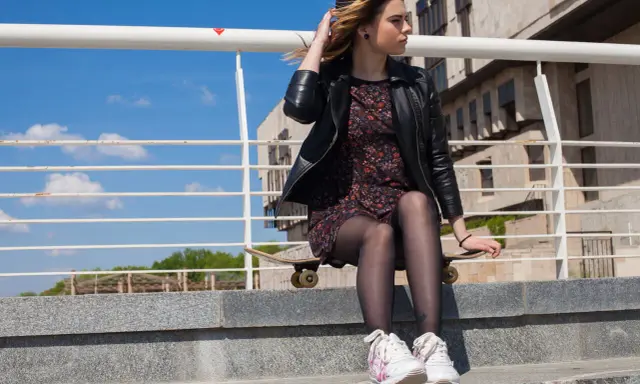 Dziewczyna w eleganckiej sukience i sportowych butach siedzi na deskorolce na schodach.