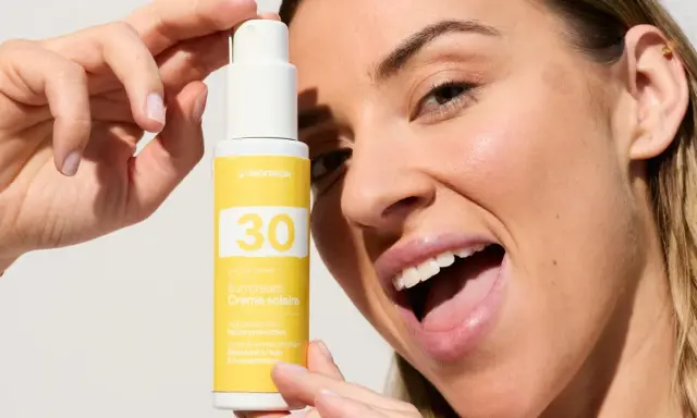 Une femme souriante tient un flacon de crème solaire visage SPF 30. La quantité de crème solaire est parfaite pour une protection optimale.