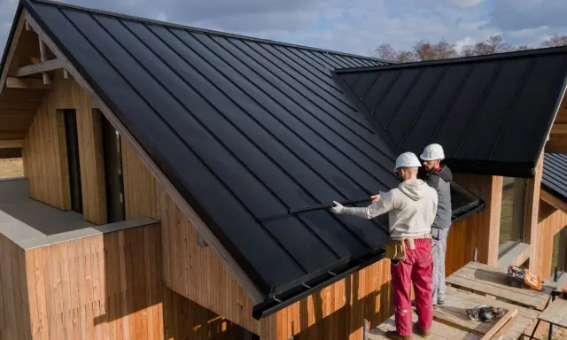Zwei Handwerker sanieren ein Dach mit schwarzer Trapezblechdeckung. Das Holzhaus erhält ein modernes Aussehen.