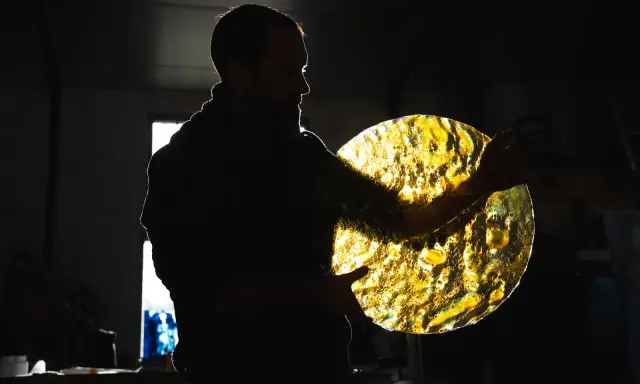 Artisan tenant un disque de verre texturé, illuminé par derrière. Les reflets dorés rappellent des vitraux.